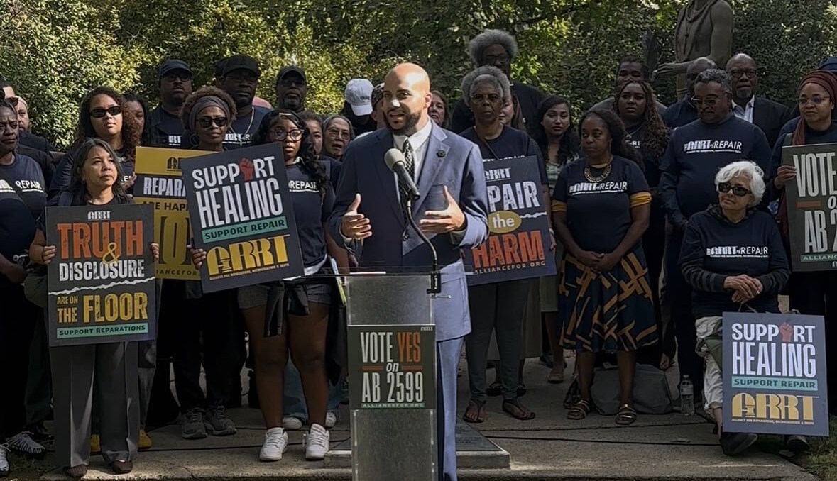 Assemblymember Isaac Bryan speaks at an outdoor podium surrounded by supporters holding signs advocating for reparations and AB 2599.